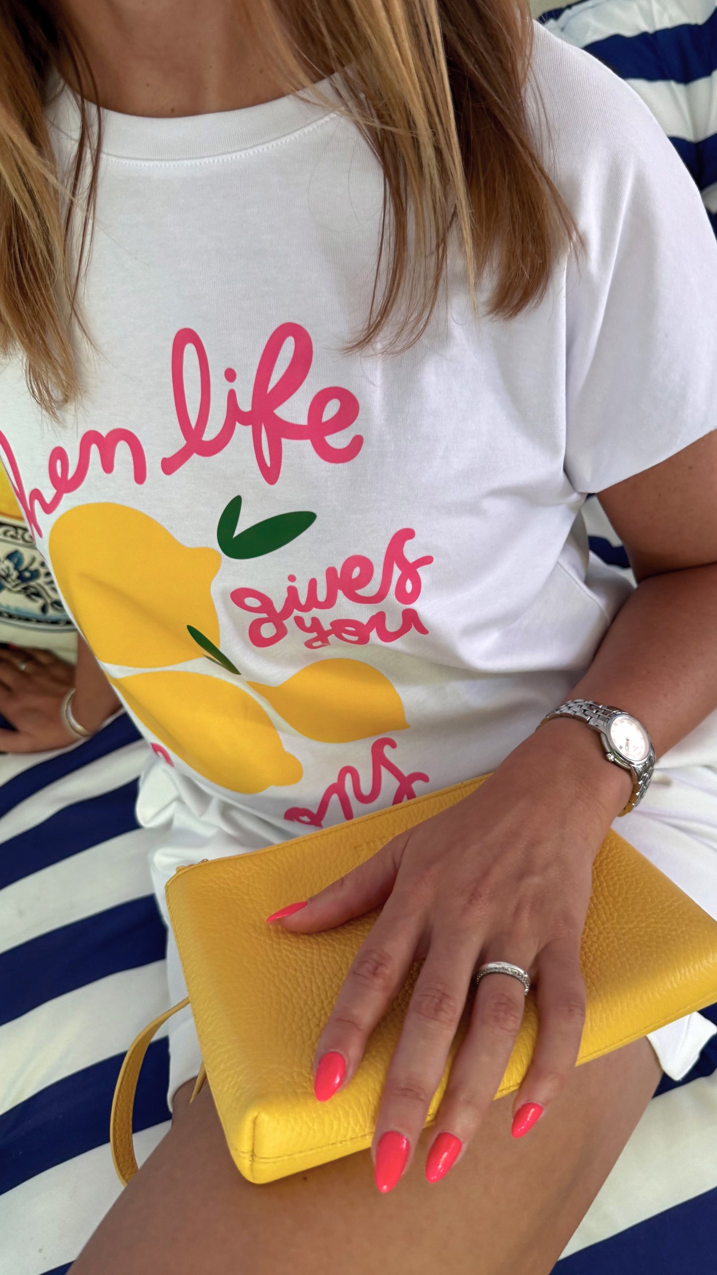Fashion Editorial in italienischem Flair Pink-Gelb- Blau: Detailzoom auf T-Shirt : Model trägt ein ausgefallenes T-Shirt mit pinker Schrift und gelben Zitronen. Sie sitzt im Urlaub im Strandcafé auf einem Sofa mit Blau Weißen Streifen.