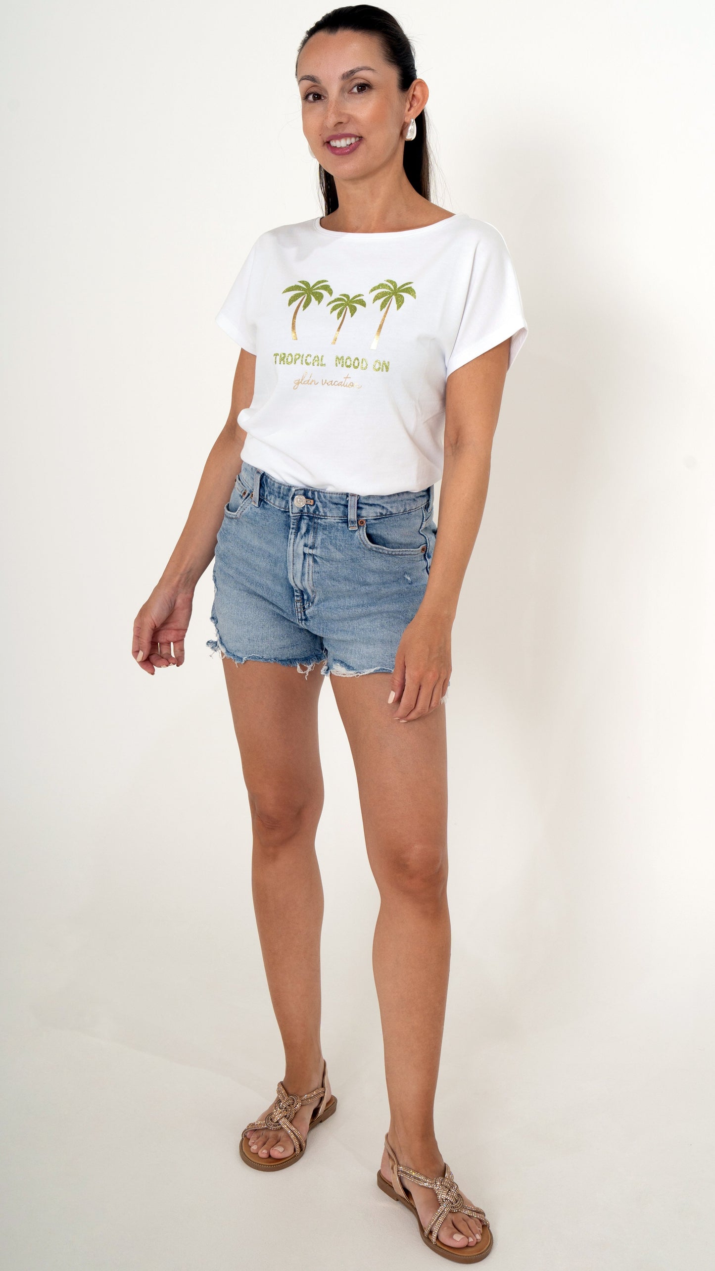 Sommerliche feminine Damenmode: Das Model trägt ein sommerliches Outfit bestehend aus einer blauen Jeans Short und ein weißes T-Shirt mit tropischem Palmenprint mit Gold Glitzer Sandalen