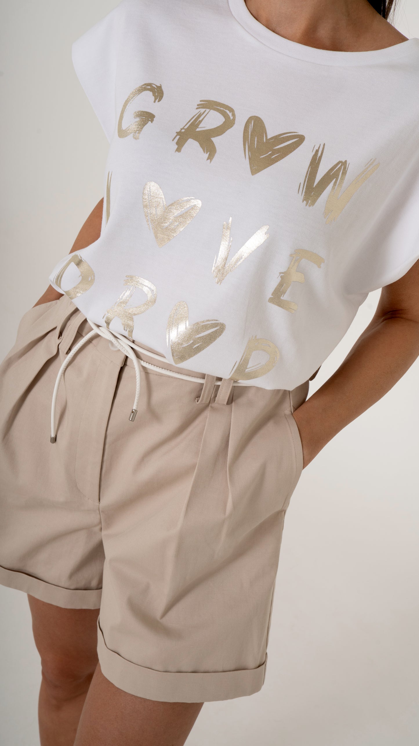 Detailansicht von sommerlich femininer Damenmode: weißes T-Shirt mit gold Glanz metallic Druck, goldene Schrift- Dazu eine beige weite Shorts mit Bundfalten und Gürtel-